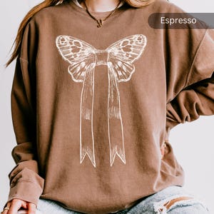 Op de afbeelding: Een bruin sweatshirt met een wit vlinderstrikontwerp. De tekst "Espresso" staat in de rechterbovenhoek van de afbeelding.