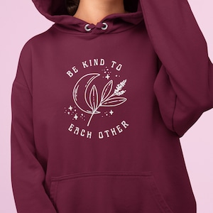 May include: A maroon hoodie featuring a white graphic with a crescent moon, leaves, stars, and the text "BE KIND TO EACH OTHER". The hoodie has a drawstring hood and a front pocket.