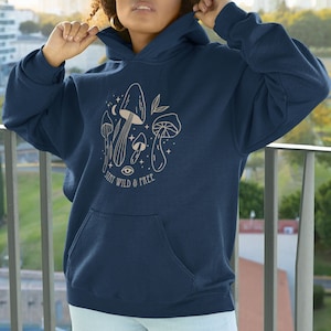 May include: Navy blue hooded sweatshirt with a front pocket and a drawstring hood. The front displays a beige illustration of mushrooms, a crescent moon, stars, and the text "STAY WILD & FREE".