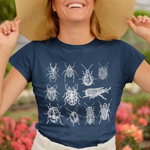 May include: A navy blue t-shirt displaying a white illustration of several insects, including beetles. The detailed graphic has a vintage, scientific style. The t-shirt is a classic crew neck design.