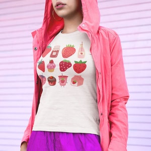 Puede incluir: Camiseta color crema con un dise&ntilde;o de fresas. El dise&ntilde;o incluye ilustraciones de fresas, mermelada de fresa, una magdalena, una bebida y un cart&oacute;n de leche de fresa. La persona lleva una chaqueta rosa y pantalones morados.