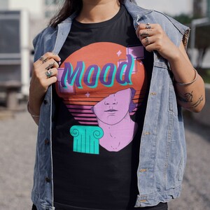May include: Black t-shirt with a retro graphic design featuring the word "Mood" in teal and purple, a bust of a statue, and a sunset-style background in orange and pink. The shirt has a classic crew neck.