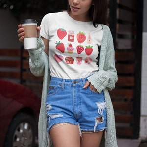 Puede incluir: Camiseta blanca con un dise&ntilde;o de fresas. El gr&aacute;fico incluye fresas, tarros de mermelada y postres de fresa. La persona lleva pantalones cortos vaqueros azules y una rebeca verde claro. La foto est&aacute; tomada al aire libre.