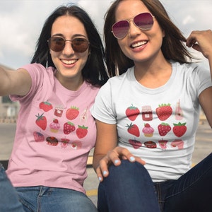 Puede incluir: Dos personas con camisetas con un dise&ntilde;o de fresas. Una es rosa, la otra blanca. Las camisetas muestran varias ilustraciones de fresas, como bayas enteras, tarros de mermelada y cupcakes.