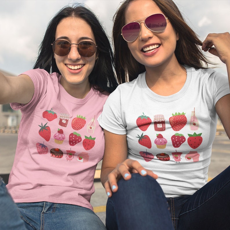 Puede incluir: Dos personas con camisetas con un dise&ntilde;o de fresas. Una es rosa, la otra blanca. Las camisetas muestran varias ilustraciones de fresas, como bayas enteras, tarros de mermelada y cupcakes.