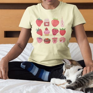 Puede incluir: Camiseta color crema con un dise&ntilde;o de fresas. El dise&ntilde;o incluye ilustraciones de fresas, mermelada de fresa, magdalenas y leche de fresa. La camiseta la lleva una persona sentada en una cama con un gato.