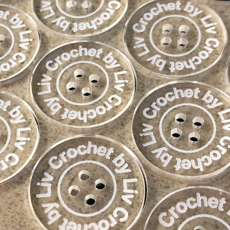 May include: Close-up of several clear plastic buttons with a brown speckled pattern. Each button has four holes and the text "Crochet by Liv" printed around the edge.