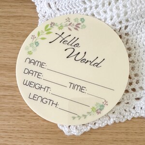 May include: A round, cream-coloured wooden plaque with a floral border and the text "Hello World". The plaque has blank lines for "Name", "Date", "Weight", "Time", and "Length".