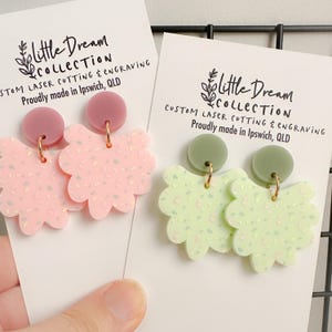 May include: Two pairs of handmade earrings with a floral design. The earrings are made of pink and green acrylic and have a speckled pattern. The earrings are attached to gold-coloured hooks.