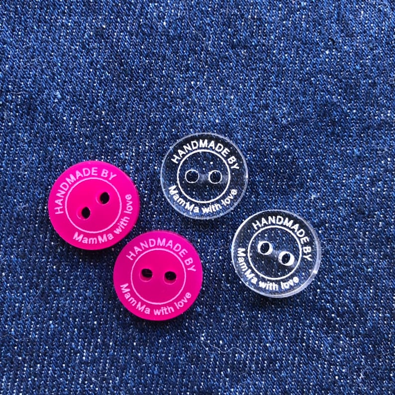 May include: Three round buttons with the text "HANDMADE BY MamMa with love" in white lettering. Two buttons are pink and one is clear.