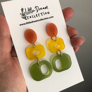 May include: A pair of dangle earrings featuring three tiers of geometric shapes. The top tier is a rounded orange shape, the middle tier is a square yellow shape with a hole in the centre, and the bottom tier is a green square shape with a hole in the centre. The earrings are displayed on a white card with the text "Little Dream Collection" and a website address.