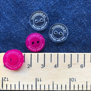 May include: Four round plastic buttons with two holes each. The buttons are approximately 1 inch in diameter. Three of the buttons are clear with white lettering that reads "HANDMADE BY MamMa with love". One button is pink with white lettering that reads "HANDMADE BY MamMa with love". The buttons are on a wooden ruler with inch markings.
