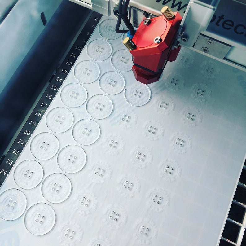 May include: A close-up of a white surface with a grid of circular cutouts. A red laser head is positioned over the surface, likely for engraving or cutting.