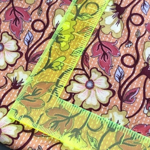 May include: Close-up of a peach-coloured fabric with a floral pattern. The fabric features a repeating design of white flowers with pink centres, brown vines, and green leaves. A yellow ruler is placed on top of the fabric.