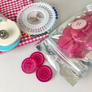 May include: A close-up of a bag of pink buttons with the text "Handmade by Esther Peake" on each button. The buttons are in a clear plastic bag with a silver zipper.