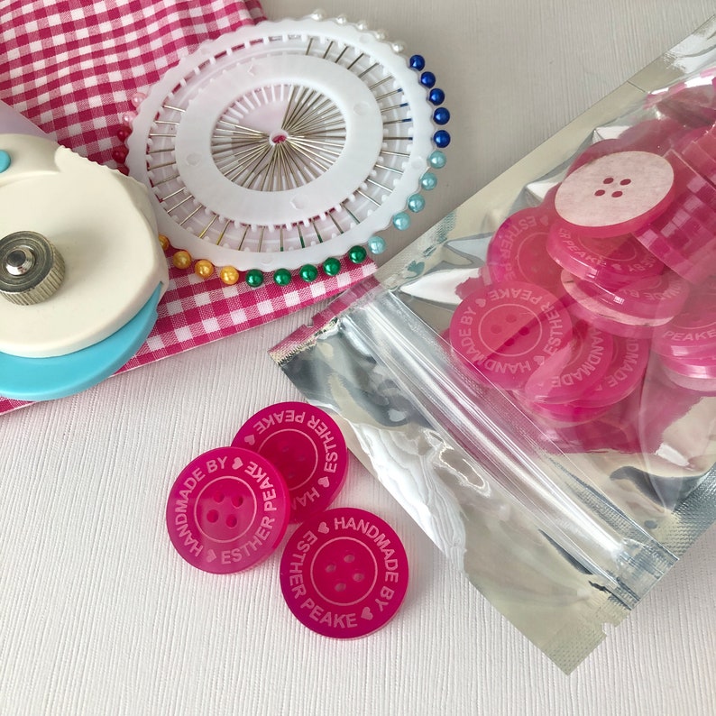 May include: A close-up of a bag of pink buttons with the text "Handmade by Esther Peake" on each button. The buttons are in a clear plastic bag with a silver zipper.