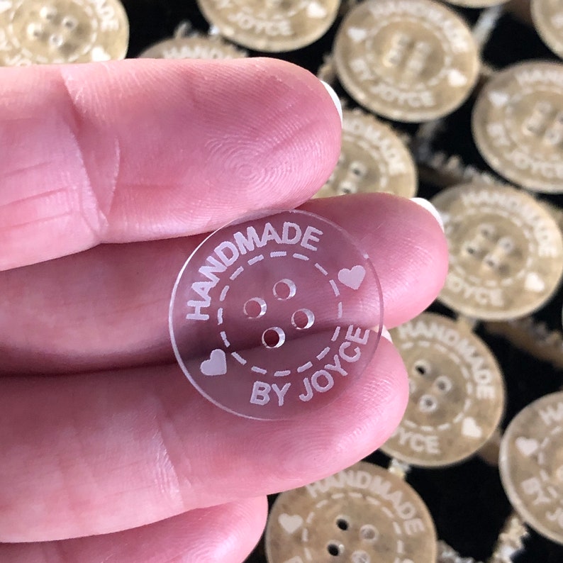 May include: Clear acrylic button with the text "HANDMADE BY JOYCE" and three holes in the center. The button is surrounded by a dashed line and two hearts.