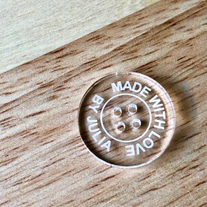 May include: Clear acrylic button with the text "MADE WITH LOVE BY JULIA" around the edge and four holes in the center.