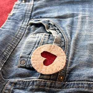 以下が含まれることがあります： 薄い青色のデニムのポケットに、茶色のフェルトでできたハート型のワッペンが貼られており、その中に赤いハートが入っています。
