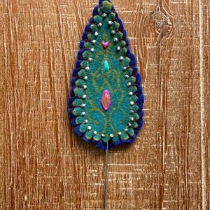 May include: A handcrafted, teardrop-shaped lapel pin. The pin features a teal and gold design with a blue border, accented with small silver beads and pink and turquoise embellishments. The pin is attached to a silver-colored metal stick pin.