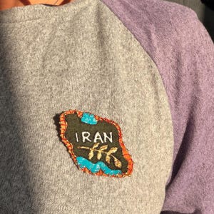 Puede incluir: Camiseta gris con mangas raglán y una manga morada, con un parche decorativo. El parche es verde oscuro con la palabra "IRAN" en blanco, detalles de hojas doradas y ribetes azules y naranjas.