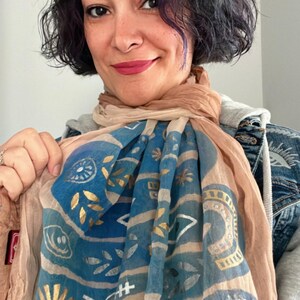 Puede incluir: Una bufanda beige y azul con detalles dorados. La bufanda presenta un patrón repetitivo de formas y símbolos geométricos. La bufanda está drapeada sobre los hombros de una persona. La persona sonríe y mira a la cámara.