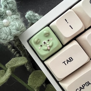 May include: A green and white custom keyboard keycap with a cartoon bear face design. The keycap is on a white keyboard with keys labeled "TAB" and "CAPSLOCK".