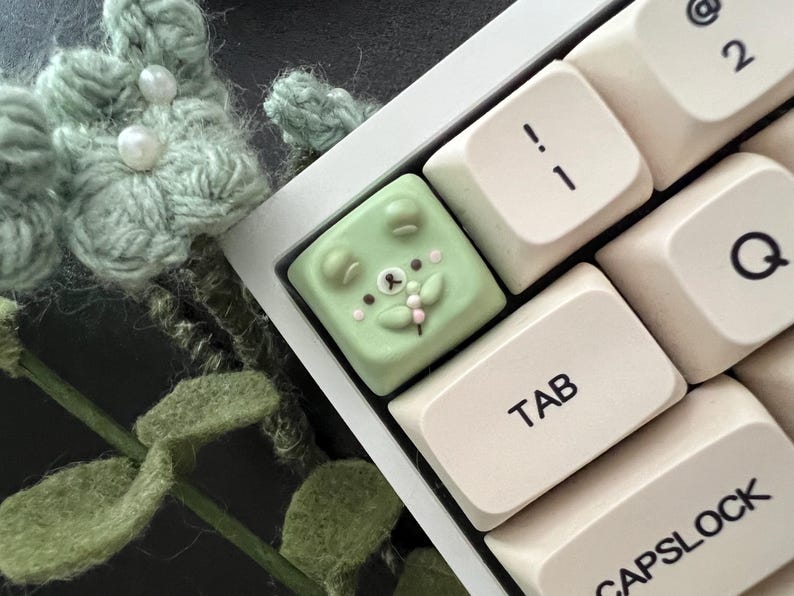 May include: A green and white custom keyboard keycap with a cartoon bear face design. The keycap is on a white keyboard with keys labeled "TAB" and "CAPSLOCK".