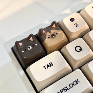 Puede incluir: Dos teclas de teclado personalizadas con caras de animales. Las teclas son marrones y tostadas con ojos negros y narices rosas. Las teclas están en un teclado blanco con las teclas "TAB" y "BLOQ MAYÚS" visibles.