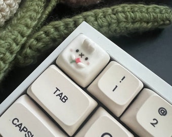 May include: A white keyboard with a custom keycap featuring a cartoon bunny with a pink nose and red cheeks. The keycap is labeled with an "X" and is located above the "TAB" key. The keyboard also has keys labeled "CAPSLOCK", "TAB", "Q", "W", "E", "R", "T", "Y", "U", "I", "O", "P", "[