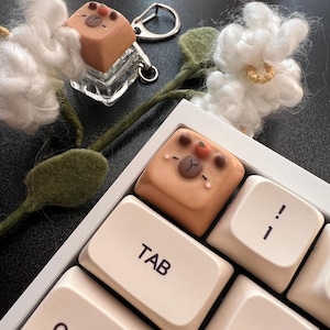 Op de afbeelding: Een close-up van een toetsenbord met een aangepaste keycap met een berenontwerp. De keycap is lichtbruin met een berengezicht en kleine oranje accenten. Een bijpassende beren keycap is bevestigd aan een sleutelhanger. Decoratieve witte katoenen bloemen en groene stelen zijn ook aanwezig.
