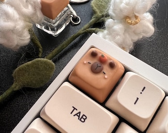 May include: A close-up of a keyboard with a custom keycap featuring a bear design. The keycap is light brown with a bear face and small orange accents. A matching bear keycap is attached to a keychain. Decorative white cotton flowers and green stems are also present.