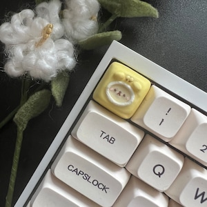 May include: A yellow keycap with a cartoon face and the text "OKS" on it. The keycap is on a white keyboard with keys labeled "TAB", "CAPSLK", "SHIFT", "Q", "W", "A", "S", "1", "2", and "!"