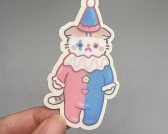 May include: A sticker of a cartoon cat dressed as a clown. The cat is wearing a pink and blue jumpsuit with a white collar and a blue and white hat. The cat has a sad expression and one blue tear on its face.