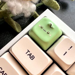 May include: A green keyboard keycap with a cute cartoon face and the word "TAB" printed on it. The keycap is on a beige keyboard.