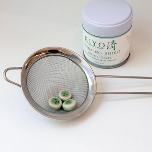 Op de afbeelding: Een fijnmazige roestvrijstalen zeef met drie kleine, ronde, lichtgroene keramische kralen met donkergroene spikkels, geplaatst naast een wit cilindrisch blik met het label KIYO CHIBA NO SHIRO Classic Grade Organic Ceremonial Matcha 40g.