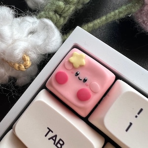 May include: A pink keycap with a cute cartoon face and a yellow star on top. The keycap is labeled "TAB" and is on a white keyboard.