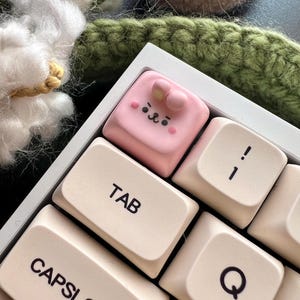 May include: A pink keycap with a cute cartoon face and a bow on top. The keycap is on a white keyboard with beige keys labeled "TAB", "CAPSLOCK", "Q", and "!".