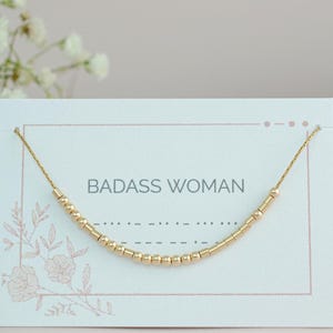 May include: A gold beaded necklace on a white card with the text "BADASS WOMAN".