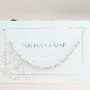 May include: A silver necklace with a Morse code design is displayed on a white card. The card has the phrase "FOR FUCK'S SAKE" printed in black. The background is a soft pink.
