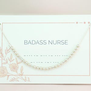 May include: A white card with a floral design and the text "BADASS NURSE" printed on it. A silver beaded necklace is displayed on the card.