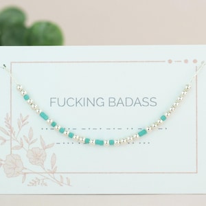 Op de afbeelding: Een delicate ketting met zilveren kralen en turquoise rechthoekige kralen wordt getoond op een witte kaart. De kaart heeft de tekst "FUCKING BADASS" in een schreefloos lettertype. Bloemenontwerpen accentueren de kaart.