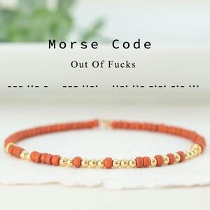 Op de afbeelding: Een kralenarmband met een morsecodebericht. De armband heeft kleine, roestkleurige kralen afgewisseld met goudkleurige kralen. De tekst boven de armband luidt "Morse Code Out Of Fucks".