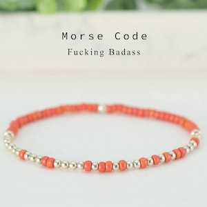 Op de afbeelding: Een armband met de tekst "Morse Code Fucking Badass". De armband heeft kleine, ronde, koraalkleurige kralen en zilverkleurige kralen. De armband ligt op een witte ondergrond met een vage groene achtergrond.