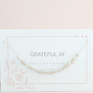 May include: A silver beaded necklace on a fine chain, presented on a white card. The card has the text "GRATEFUL AF" and a floral pattern. The necklace is a minimalist, stylish accessory.