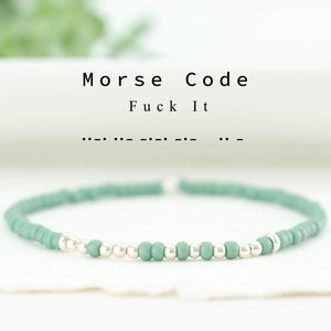 May include: A beaded bracelet with teal and silver beads. The bracelet is displayed on a white surface, with the words "Morse Code Fuck It" above it. The Morse code for "Fuck It" is also shown.