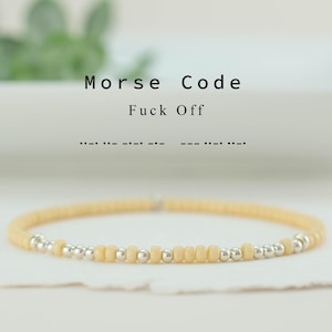 Op de afbeelding: Een delicate armband met kleine, lichtgele kralen en zilveren accenten. De tekst "Morse Code Fuck Off" staat boven de armband. De Morsecode voor "Fuck Off" wordt ook getoond. De armband ligt op een wit oppervlak.