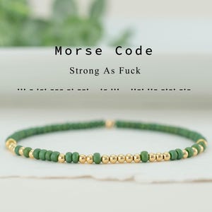 May include: A green beaded bracelet with gold accents. The bracelet features small green beads interspersed with gold beads. The text "Morse Code Strong As Fuck" is above the bracelet.