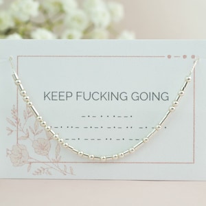 以下が含まれることがあります： 白いカードに花柄のデザインが施され、"KEEP FUCKING GOING"というテキストが書かれた、銀色のビーズネックレスです。