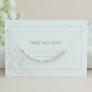 Op de afbeelding: Een delicate zilveren ketting met een Morsecode-ontwerp dat "TAKE NO SHIT" spelt, wordt getoond op een witte kaart. De kaart heeft een roze rand en bloemaccenten. De achtergrond bevat zachte roze en witte elementen.